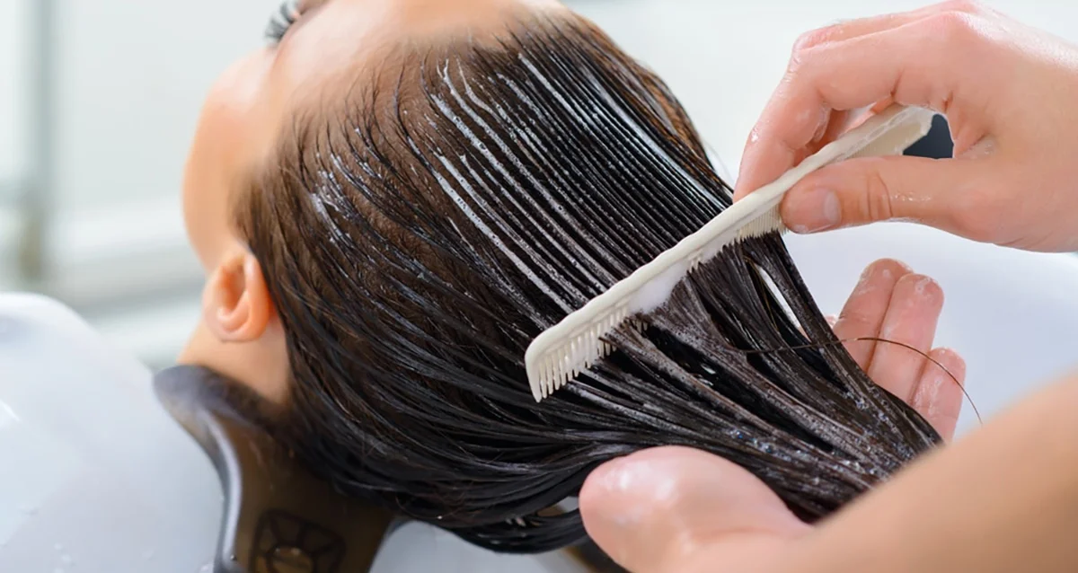 Hands applying hair treatment using a comb on wet hair at a salon sink.